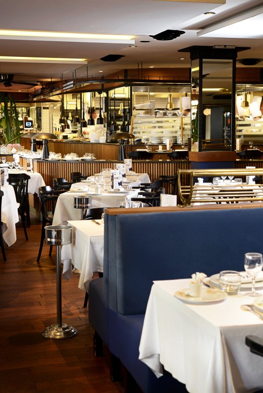 Vue intérieure de la salle de la Brasserie L’Est à Lyon, avec tables élégamment dressées et cuisine ouverte en arrière-plan.
