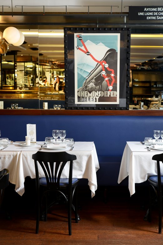 Salle de la Brasserie L’Est à Lyon avec banquette bleue, tables dressées et affiche vintage de train.