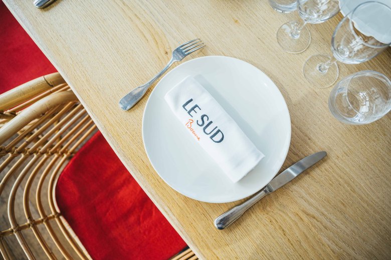 Assiette blanche avec serviette brodée Le Sud posée sur une table en bois clair à la Brasserie Le Sud à Lyon.